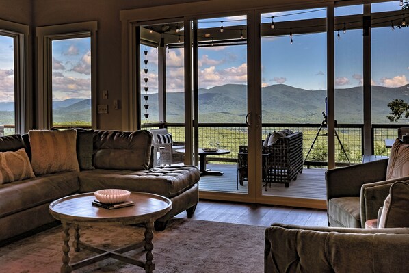 A spacious living room with a large sliding glass door leading to a deck, a leather sofa, a round coffee table, and a view of mountains.