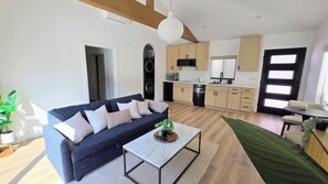 Ein modernes Wohnzimmer mit einem dunklen Sofa, einem kleinen Couchtisch und einer Küchenzeile mit hellen Holzschränken und einem schwarzen Backofen.