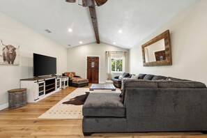 Uma sala de estar com sofá cinzento, piso de madeira, televisão e teto rústico.