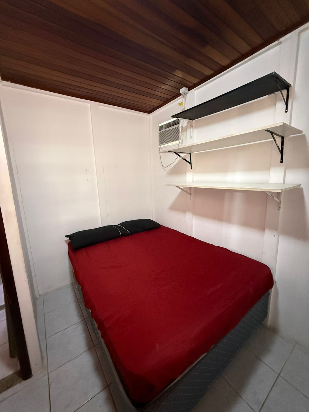 A small room with a single bed, white walls, a wooden ceiling, and a shelf unit with an air conditioning unit.
