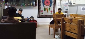 Una habitación con un banco de madera, una guitarra y una persona tocándola. Hay un tapiz colorido en la pared y un estante con botellas.