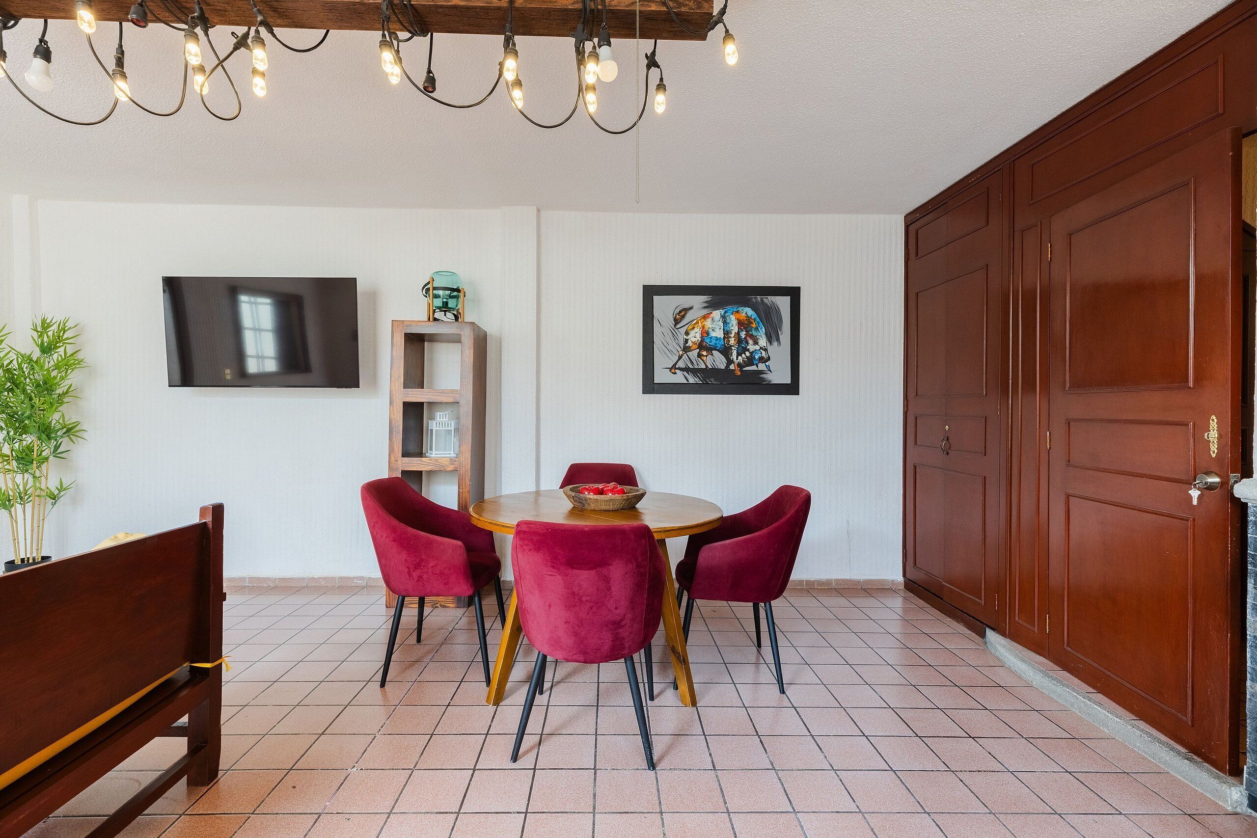 Un coin repas comprenant une table ronde, quatre chaises, un banc, un téléviseur, une étagère et une porte.