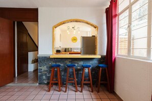 Una cocina con encimera de piedra, taburetes de madera, un refrigerador y una ventana con cortina.