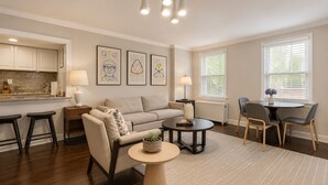 Un salon moderne avec un canapé, une table basse et des fauteuils, un coin cuisine doté de placards et d’un plan de travail, et des œuvres d’art accrochées aux murs.