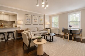 Ein modernes Wohnzimmer mit einer Couch, einem Couchtisch und Sesseln, einem Küchenbereich mit Schränken und einer Arbeitsplatte sowie Kunstwerken an den Wänden.