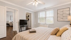 Une chambre à coucher comprenant un lit, un téléviseur, un ventilateur de plafond, une fenêtre avec des stores et une porte donnant sur une autre pièce.
