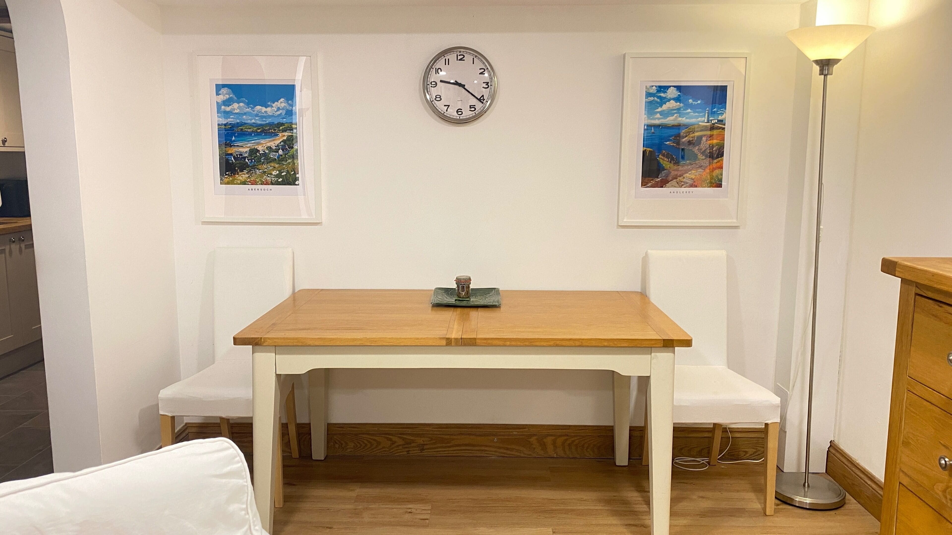 Ein Zimmer mit einem Holztisch, zwei weißen Stühlen, einer Uhr und zwei gerahmten Bildern an der Wand.