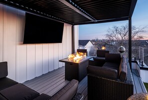 Une terrasse extérieure dotée d’un téléviseur à écran plat, d’un foyer, de meubles en osier et offrant une vue sur le paysage urbain.