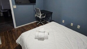 Quarto de hotel com uma cama, uma escrivaninha, uma cadeira, uma TV fixada na parede e uma porta que dá para outro cômodo.