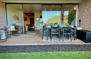 A modern patio with a dining table and chairs, a sofa, and a large window overlooking a garden. - Surf Rider, Newly Remodeled, Walk Out Lanai, Ocean View, 2ACs, 350Steps to Kam 1 (Kihei)