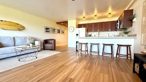 A modern living room with a sofa, coffee table, and a dining area with bar stools. - Surf Rider, Newly Remodeled, Walk Out Lanai, Ocean View, 2ACs, 350Steps to Kam 1 (Kihei)