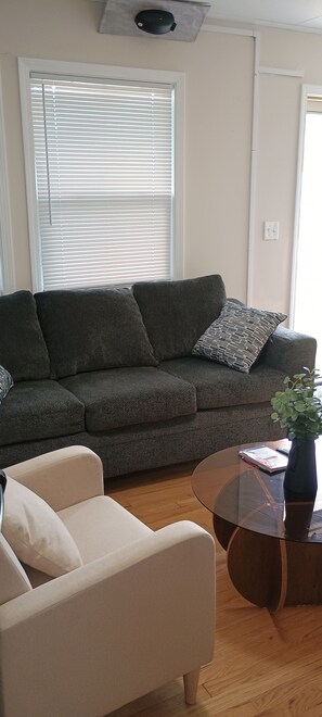 Een woonkamer met een donkergrijze bank, een lichtbeige fauteuil en een glazen salontafel met een boek en een plant.