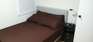 Een bed met een bruine spreidek, een nachtkastje met een lamp en een raam met zonwering.