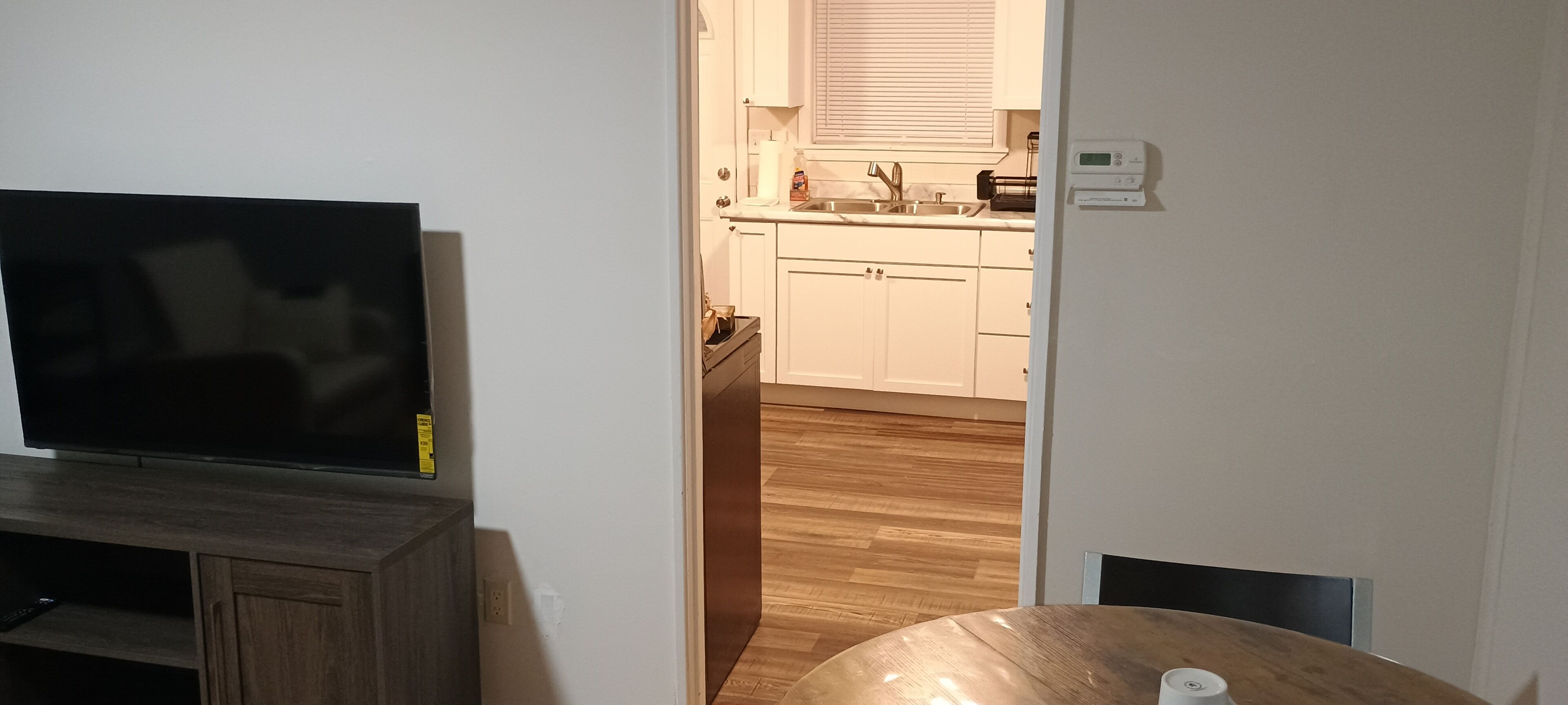 Ein Zimmer mit Fernseher auf einem Holzständer, einem Esstisch mit einer weißen Tasse und einer durchgehenden Tür sichtbaren Küche.