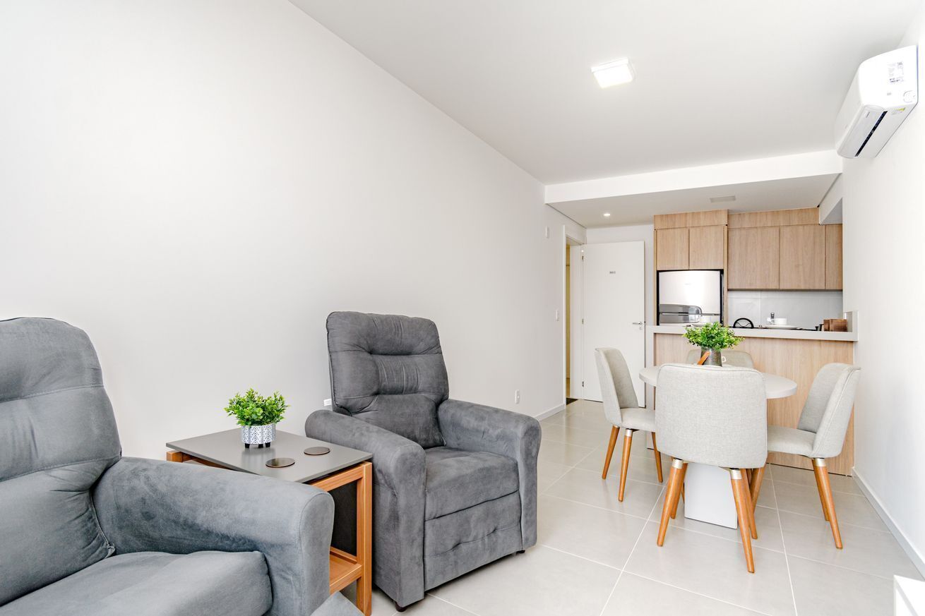 A modern living room with grey sofas, a small table with a plant, and a dining area with a white table and grey chairs.