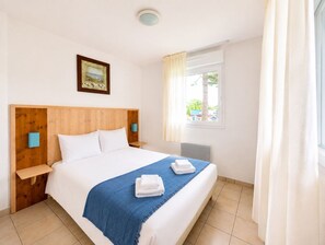 Une chambre d’hôtel avec une tête de lit en bois, un lit recouvert de draps blancs, un plaid bleu et une fenêtre avec des rideaux.