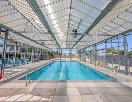 A covered swimming pool with clear blue water, surrounded by chairs and a glass ceiling structure.. L'Escale de l'Estuaire, T3 with Pool Near Soulac
