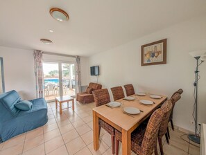 A dining area with a table set for four, a blue sofa, wicker chairs, a TV mounted on the wall, and a view through a glass door. - L'Escale de l'Estuaire, T3 with Pool Near Soulac (Le Verdon-sur-Mer)