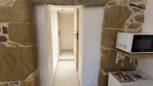 A narrow hallway with stone walls, a white door at the end, a white refrigerator with a magazine, and a small trash bin. - Small 4 person self-catering apartment in LEDENON - Entire apartment (Lédenon)