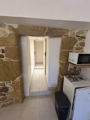 Ein schmaler Korridor mit Steinwänden, einer weißen Tür am Ende, einem weißen Kühlschrank mit einer Zeitschrift und einer kleinen Mülltonne.