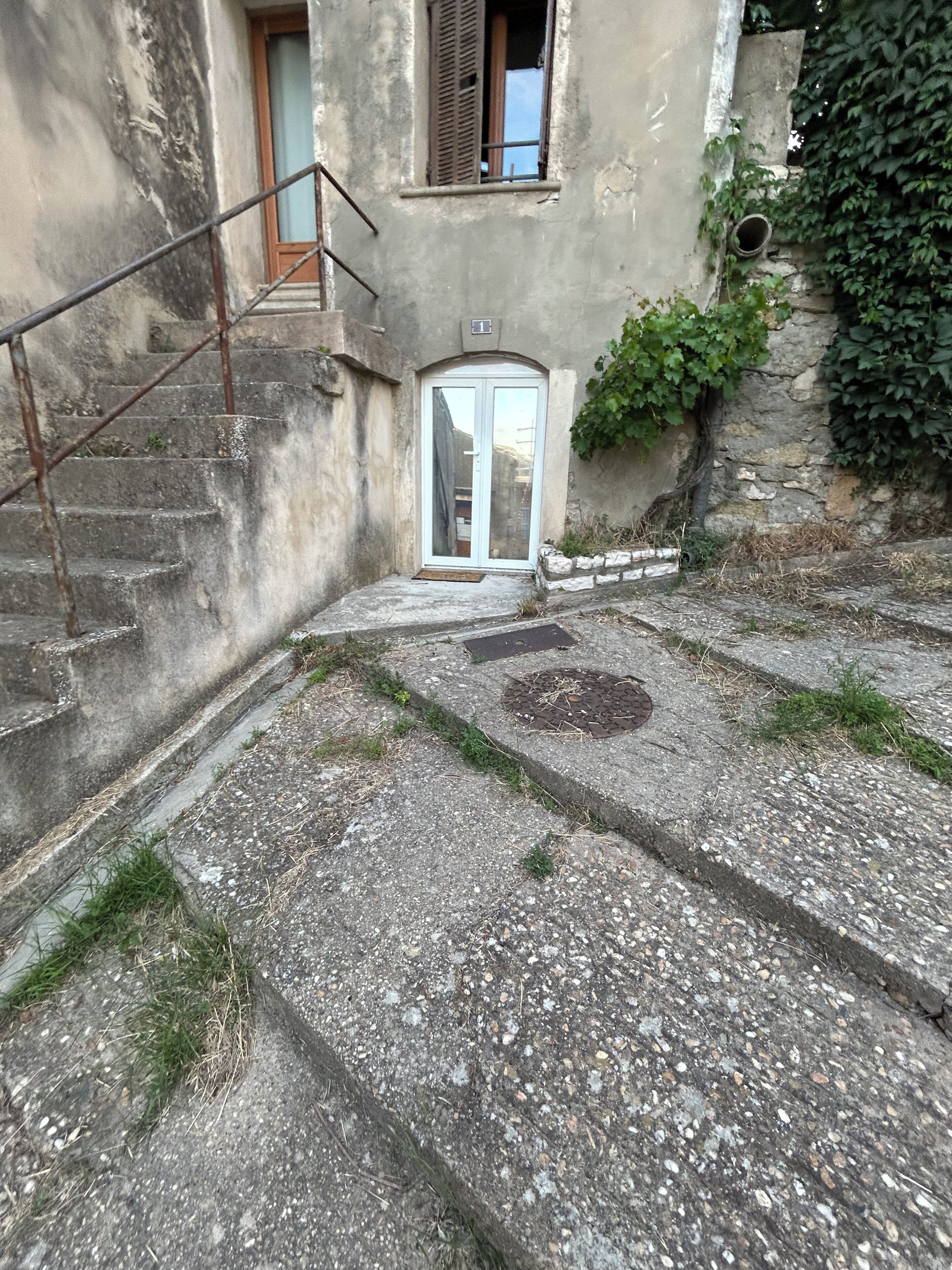 Ein Gebäude mit einer Treppe, die zu einer Tür führt, einem kleinen Fenster mit Fensterläden und einer Steinmauer mit Grünpflanzen.