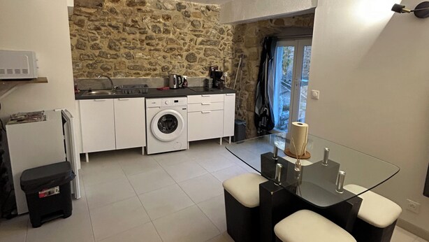 A compact kitchen with a stone wall, a washing machine, and a dining area with a glass table and black chairs. - Small 4 person self-catering apartment in LEDENON - Entire apartment (Lédenon)