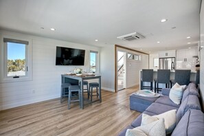 A modern living room with a sofa, a dining area with a table and chairs, a flat-screen TV mounted on the wall, and a kitchen area with cabinets and a refrigerator. - Sleeps 20 Walk to Beach 2 Side-by-Side Homes (San Diego)