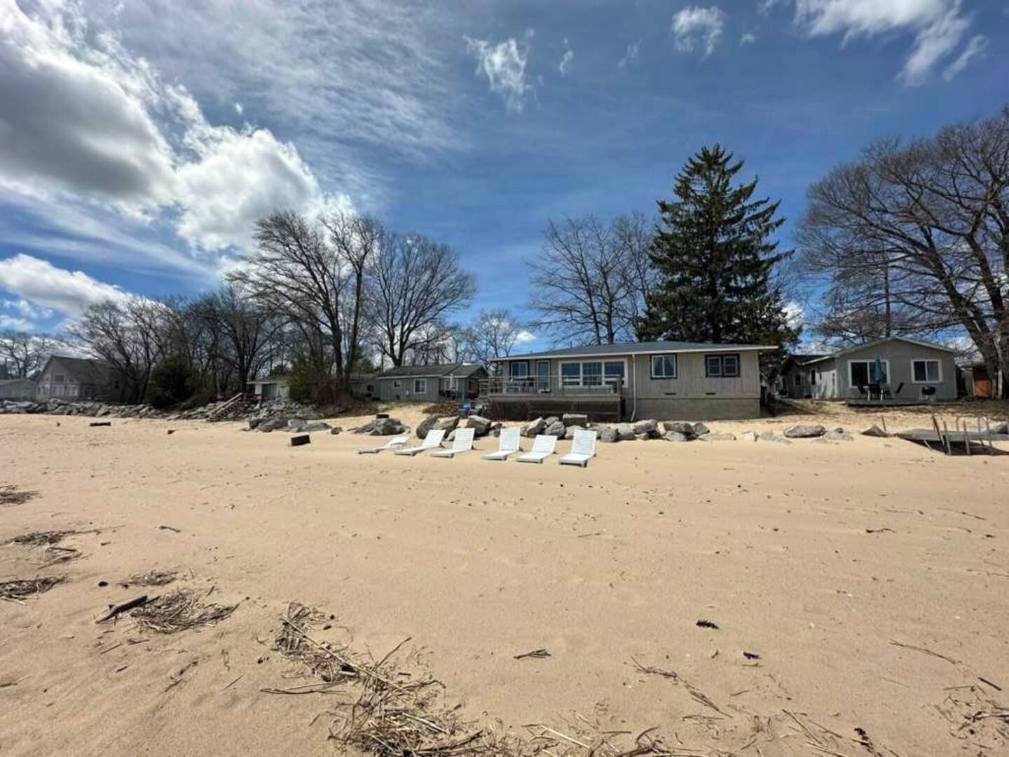 Ett hus vid stranden med en sandig trädgård framför, flera solstolar och ett stort träd.