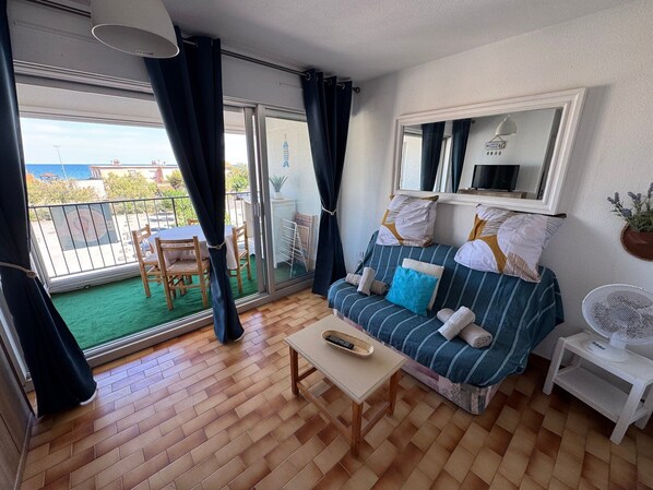 A room with a sofa, a table, a chair, a balcony with a view, and a fan. - L’Escapade du Cap, Sea View, Sleeps 6 (Le Cap d'Agde)