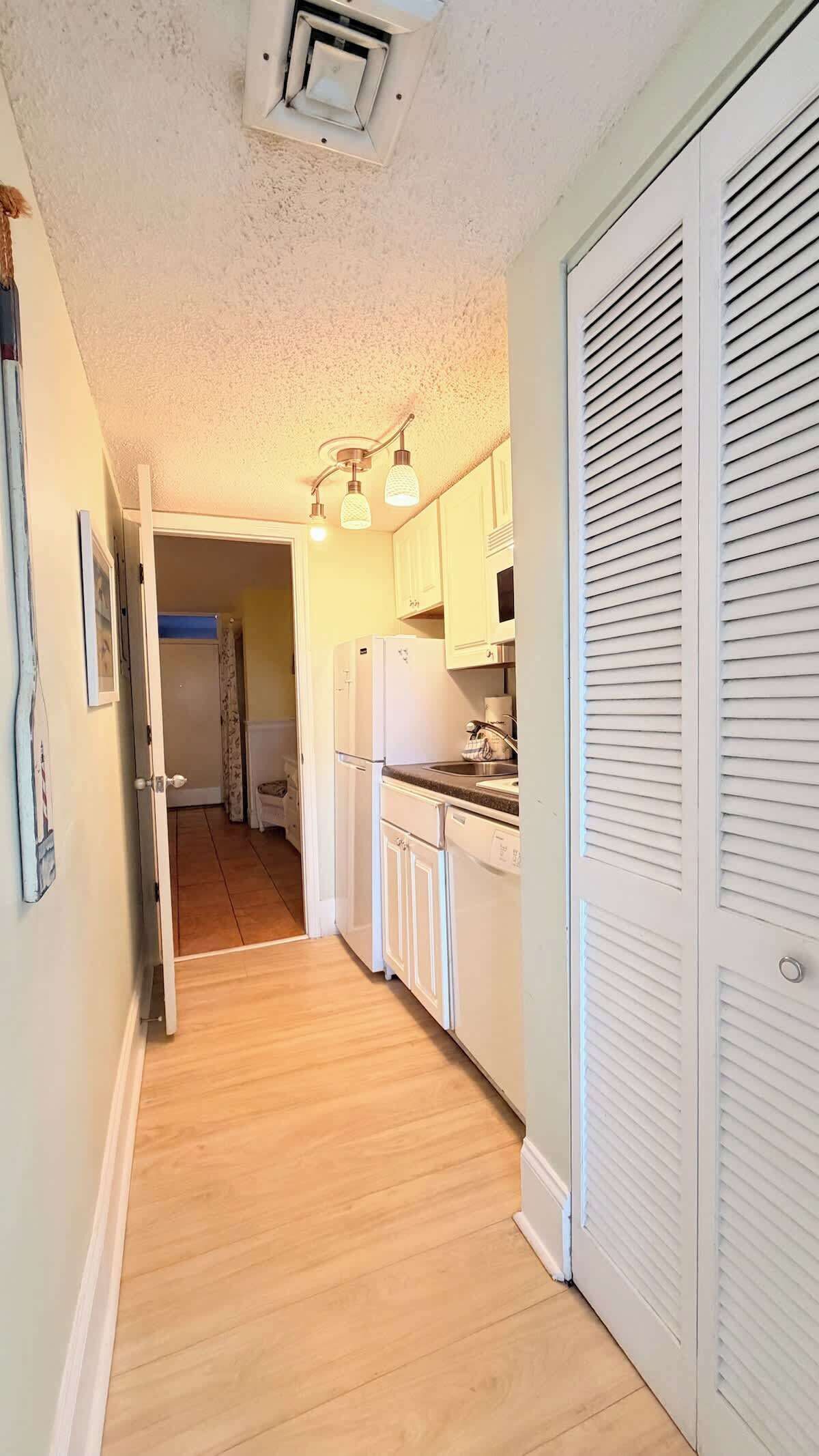 A narrow hallway with a wooden floor, a white door, and a kitchen area with white cabinets and a refrigerator.
