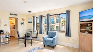 A living room with a dining area, a blue armchair, a TV, and a view of the ocean through large windows. - Beachside Condo Near Ocean | Pool | Sleeps 6 (Myrtle Beach)