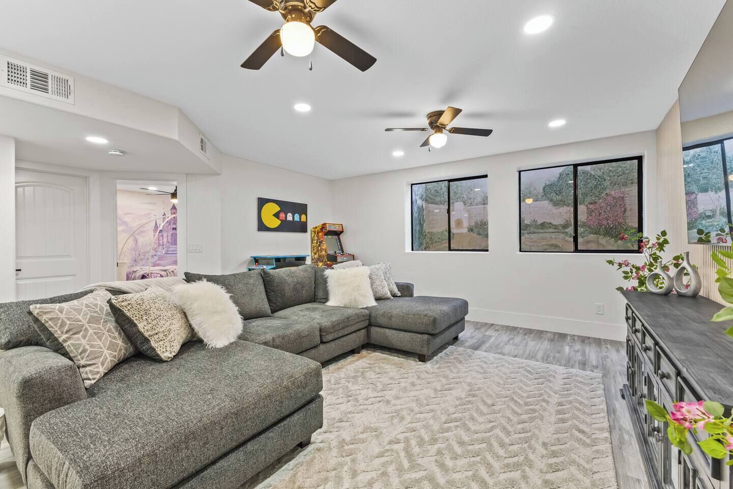 A modern living room with a sectional sofa, two ceiling fans, and large windows.