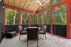 Ein überdachter Außenbereich mit rundem Tisch und Stühlen, umgeben von großen Fenstern mit Blick auf einen Wald.