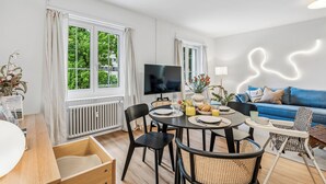 A modern living room with a dining area, large windows, a flat-screen TV, and a blue sofa.
