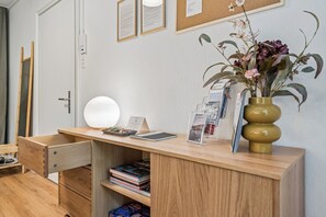 Uma escrivaninha de madeira com uma luminária, um vaso com flores e um mural com papéis emoldurados.