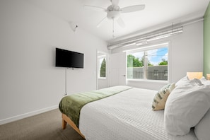A bedroom with a bed, a ceiling fan, a TV, a window, and a door.
