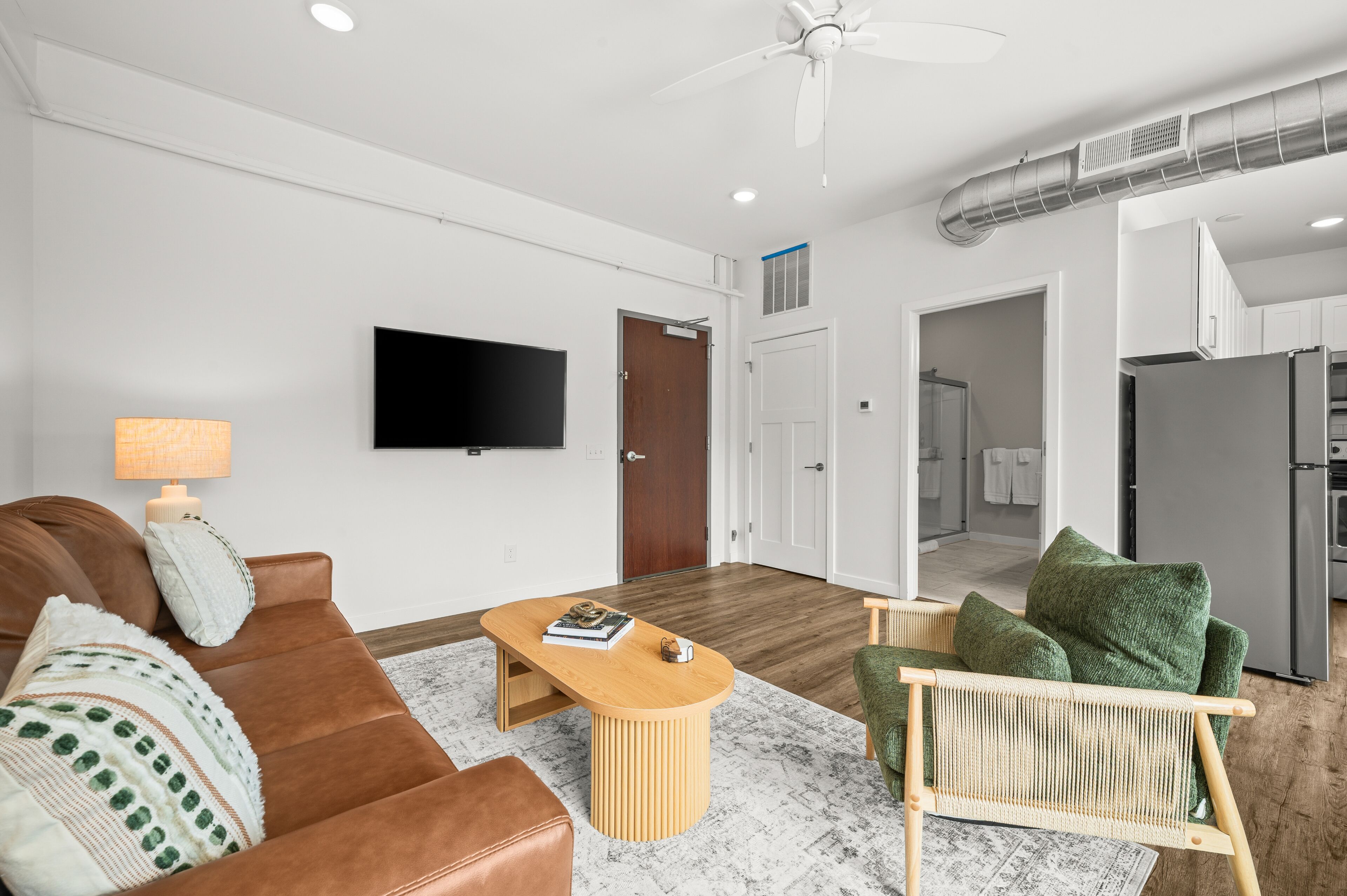 A modern living room with a flat-screen TV, a brown leather sofa, a wooden coffee table, and a green armchair.