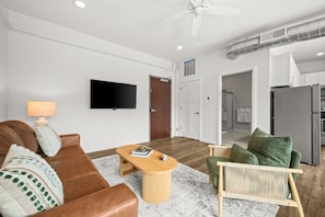 A modern living room with a flat-screen TV, a brown leather sofa, a wooden coffee table, and a green armchair.