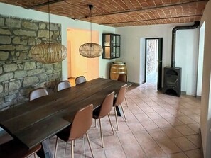Ein Essbereich mit einem langen Tisch, Stühlen, Pendelleuchten, einer Steinwand und einem Kaminofen.