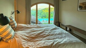 Ein Schlafzimmer mit Bett, Lampe und Blick auf ein blaues Gewässer, sowie einer Holzbalkendecke.