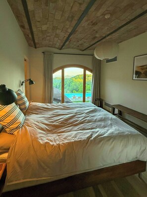 Ein Schlafzimmer mit Bett, Lampe und Blick auf ein blaues Gewässer, sowie einer Holzbalkendecke.