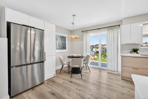 Une cuisine moderne dotée d’un réfrigérateur en acier inoxydable, d’une table à manger avec des chaises et offrant une vue sur l’extérieur grâce à de grandes fenêtres.