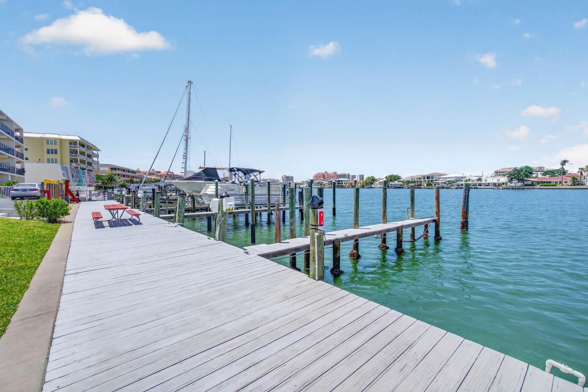 A waterfront dock with wooden planks, a marina with boats, and buildings in the background.