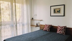 Ein Schlafzimmer mit einem Bett, zwei Kissen, einem Nachttisch, einer Lampe, einem Regal mit einem Bild und einem Fenster mit Vorhängen.