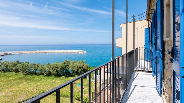 A balcony with a view of the sea and a distant pier, a stone building with blue shutters, and a clear sky.