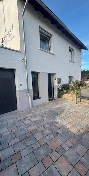 A modern two-story house with a paved driveway, a garage, and a small garden area with a bench. - Small 45-square-meter apartment in the vineyards—ideal for hiking and mountain biking (Bad Kreuznach)