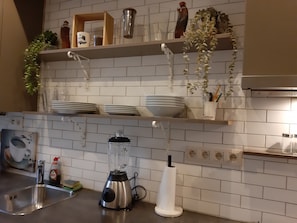 Una cocina con salpicadero de azulejos blancos, un lavamanos, una licuadora y estantes con vajilla y utensilios de cocina.