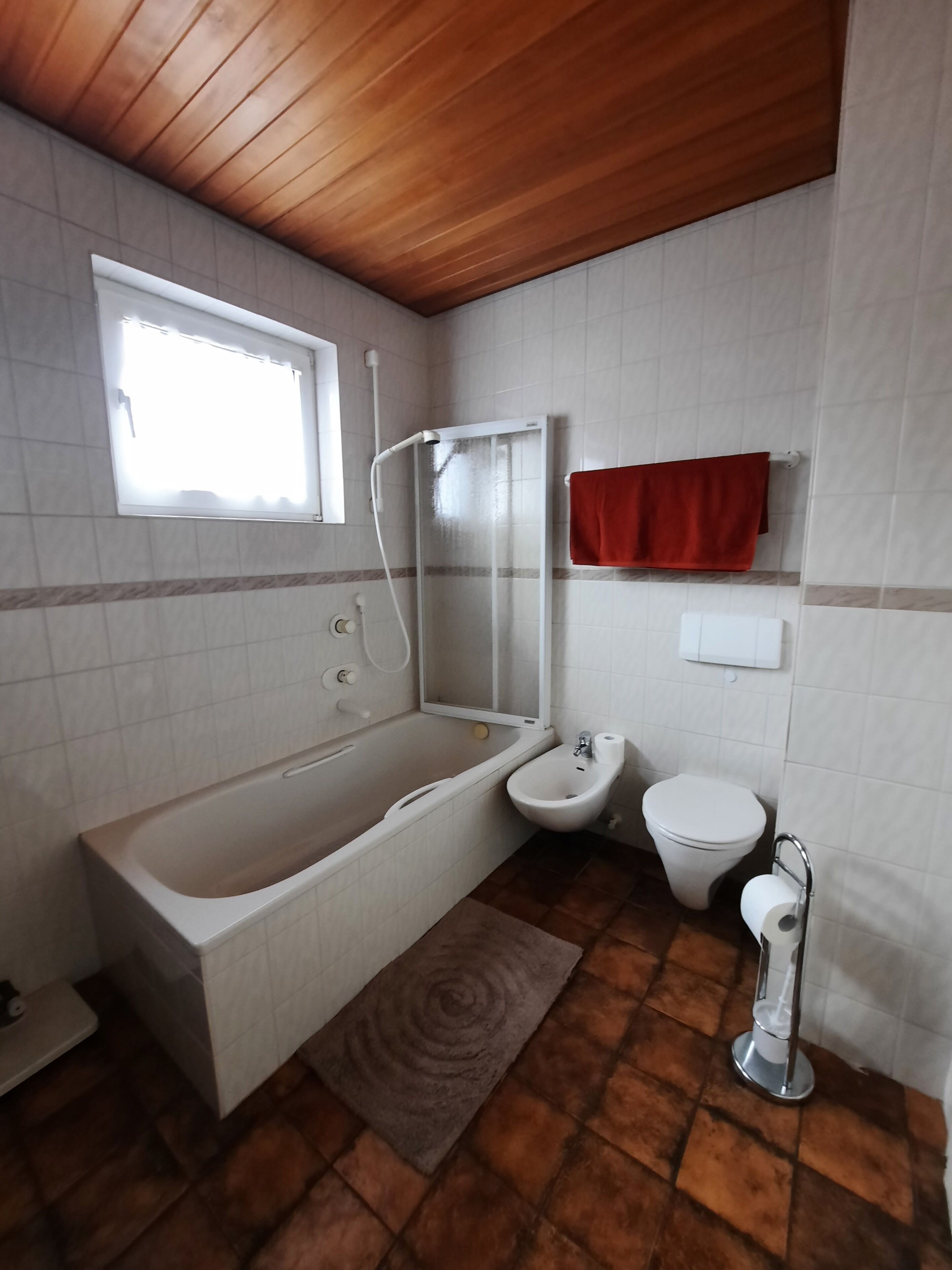 Een badkamer met een badkuip, een hygiëneschuimer, een toilet en een raam.