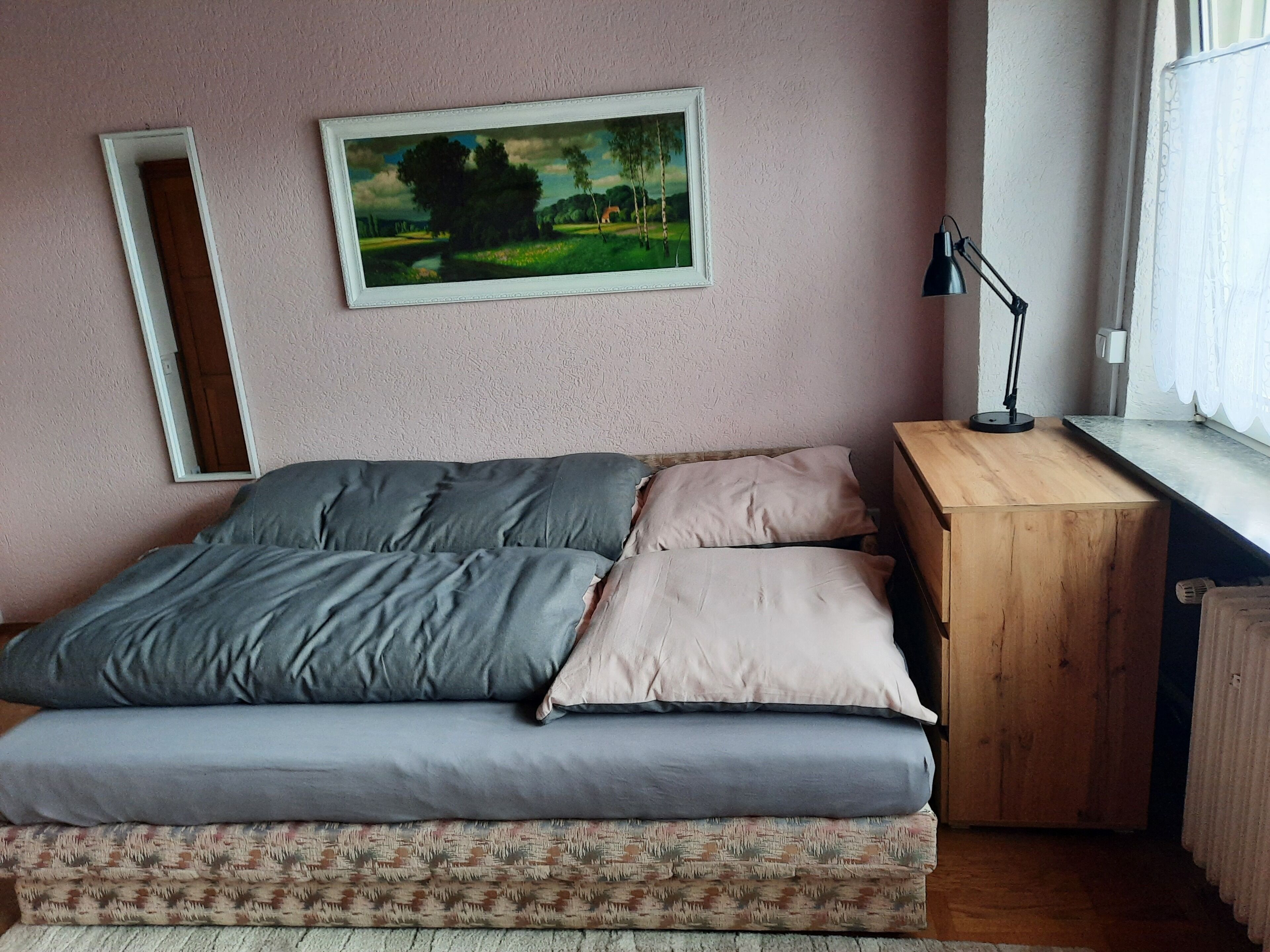 Ein Schlafzimmer mit einem Bett, einem Nachttisch aus Holz, einem Schreibtisch, einem Spiegel und einem Fenster mit Vorhängen.