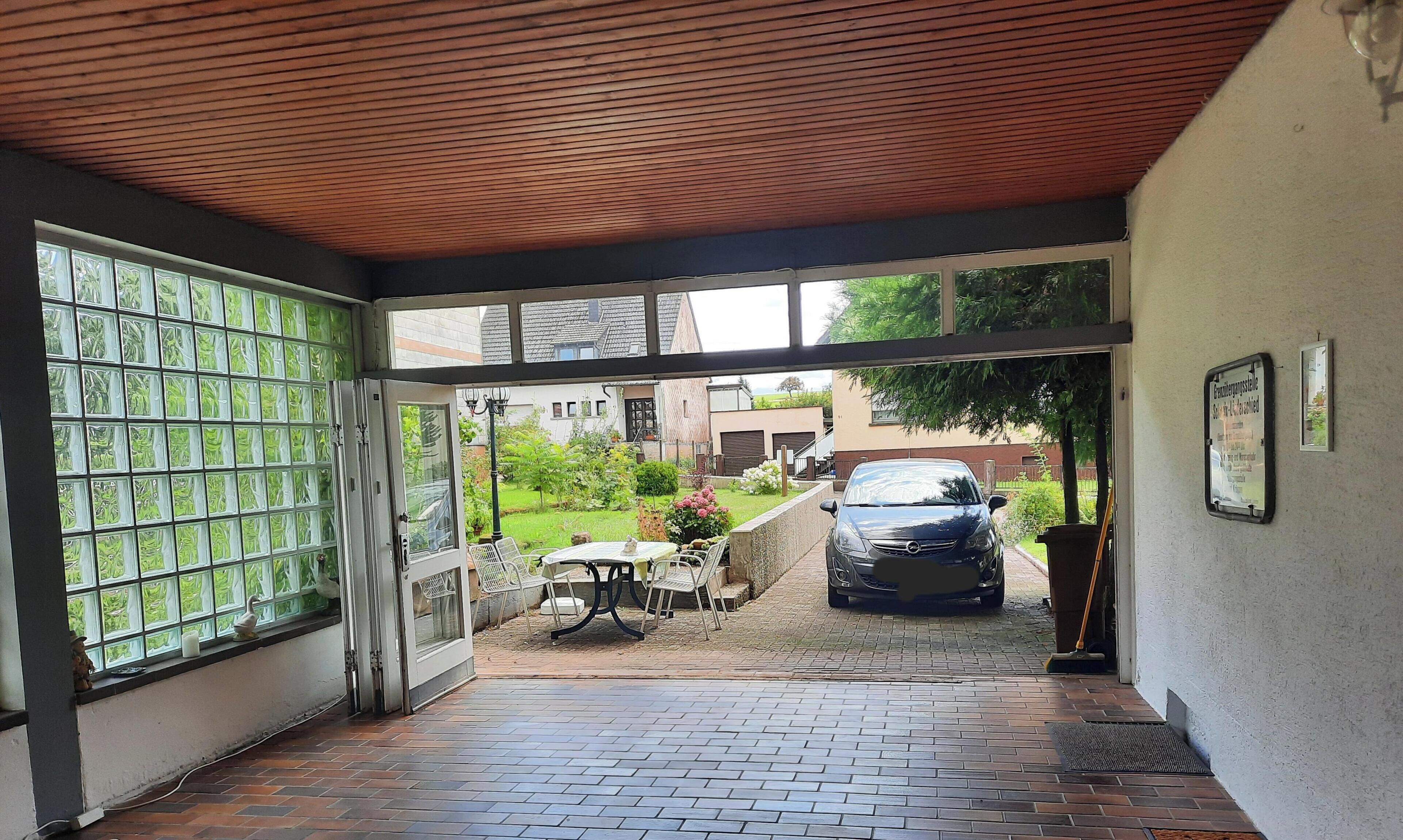 Ein überdachter Außenbereich mit einem geparkten Auto, einem Tisch mit Stühlen und Blick auf eine Wohngegend.